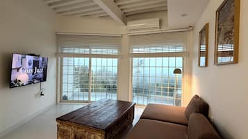 Uma sala de estar com televisão, mesa de centro de madeira, sofá marrom e uma janela grande com persianas.