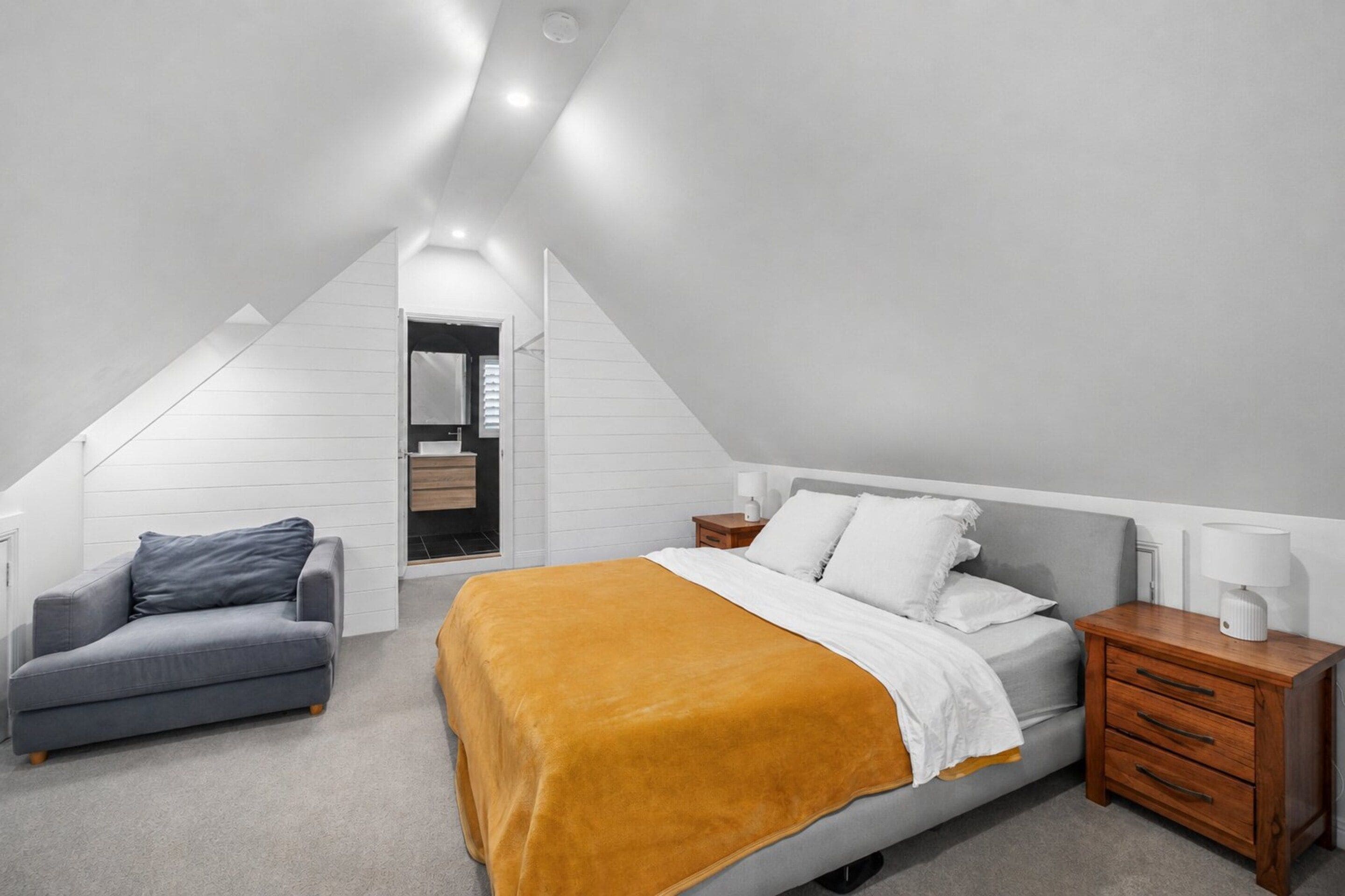A modern bedroom with a sloped ceiling, a bed with white bedding, a gray sofa, and a wooden nightstand.