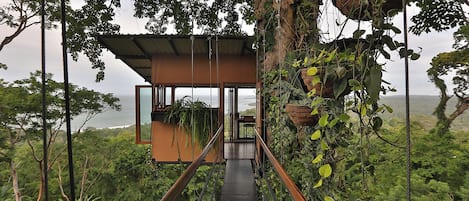 Uma casa na árvore com fachada de madeira, escada de metal e vista para o oceano.