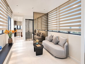 A modern living room with a sofa, a coffee table, and a vase with flowers. There is a glass partition and a view of the city through large windows with blinds.