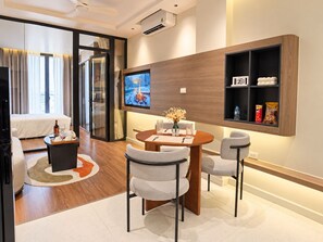 A modern hotel room with a dining area, a TV mounted on the wall, and a shelf with snacks. - An Home (Ngoc Ha)