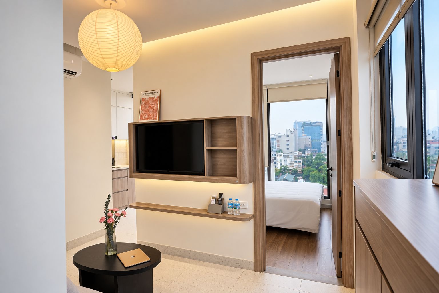 A modern hotel room with a flat-screen TV, a bedside table with a laptop, a vase with flowers, and a city view through the window.