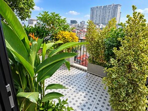 A balcony with a tiled floor, surrounded by various plants and a cityscape in the background. - An Home (Ngoc Ha)