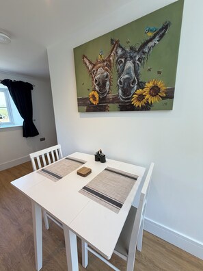 Un comedor con una mesa blanca, dos individuales tejidos y un cuadro de caballos y girasoles en la pared.