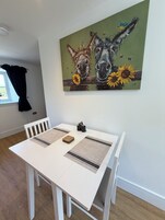 Un comedor con una mesa blanca, dos individuales tejidos y un cuadro de caballos y girasoles en la pared.