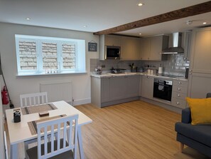 Een moderne keuken met grijze kastjes, een magnetron en een kookplaat. Er is een eettafel met stoelen en een bank met een geel kussen.
