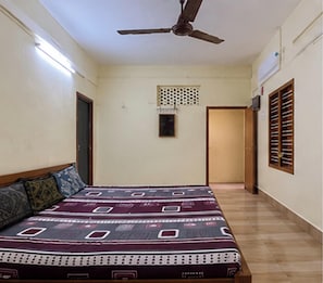 A bedroom with a bed, a ceiling fan, a door, and a window with shutters. - TMS Home Stay (Tiruchendur)