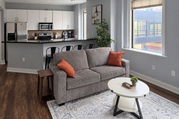 A modern living space with a grey sofa, a round coffee table, and a kitchen area with white cabinets and appliances.