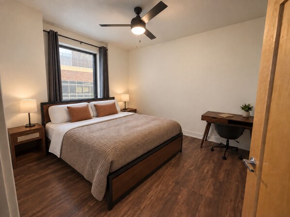 Un dormitorio con cama, mesitas de noche, escritorio, silla, ventilador de techo y ventana con cortinas.