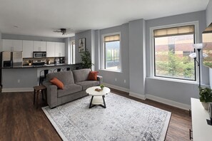A modern living room with a grey sofa, a round coffee table, and a kitchen in the background. - The City Escape - Skywalk + Dining (Des Moines)