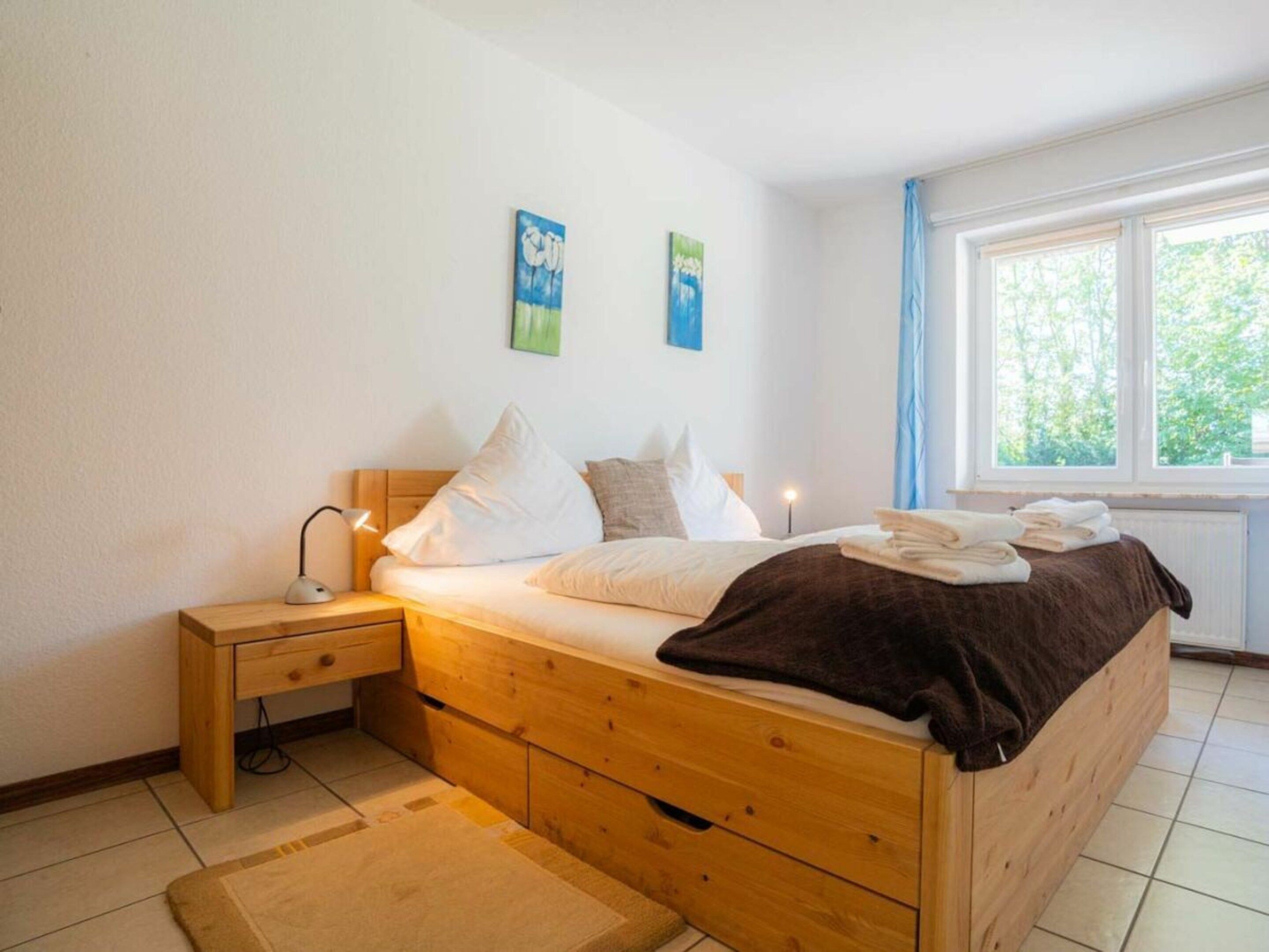 Quarto com cama de madeira, mesa de cabeceira, abajur e duas pinturas na parede.
