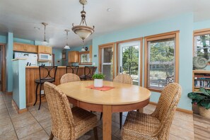 A dining area with a round table, wicker chairs, a central plant, and a kitchen in the background. - The Little Mermaid - Dogs OK - Minutes to beach (Pacific City)