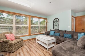 A living room with a sofa, armchair, and coffee table. There are large windows with blinds, a door, and a wall-mounted light fixture. - The Little Mermaid - Dogs OK - Minutes to beach (Pacific City)