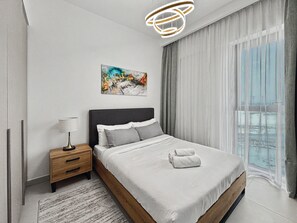 Ein modernes Schlafzimmer mit einem großen Bett, einem Nachttisch, einer Lampe, einem Gemälde und Blick ins Freie.
