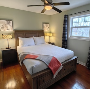 A bedroom with a bed, two bedside tables, a ceiling fan, a window with curtains, and artwork on the walls. - L&V | Victorian Garden Retreat | 3BR| Spacious Deck | Walk to Historic Old Town (Niagara-on-the-Lake)