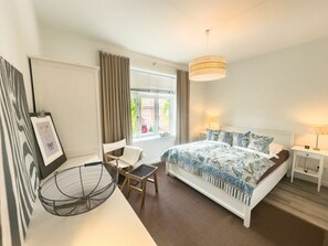 Ein Hotelzimmer mit Bett, Sessel, Schreibtisch, Lampe und einem fenster mit Vorhängen.