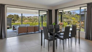 Et moderne spiseområde med en glasdør, der fører til en udendørs terrasse, et spisebord med stole og udsigt til en anlagt have.