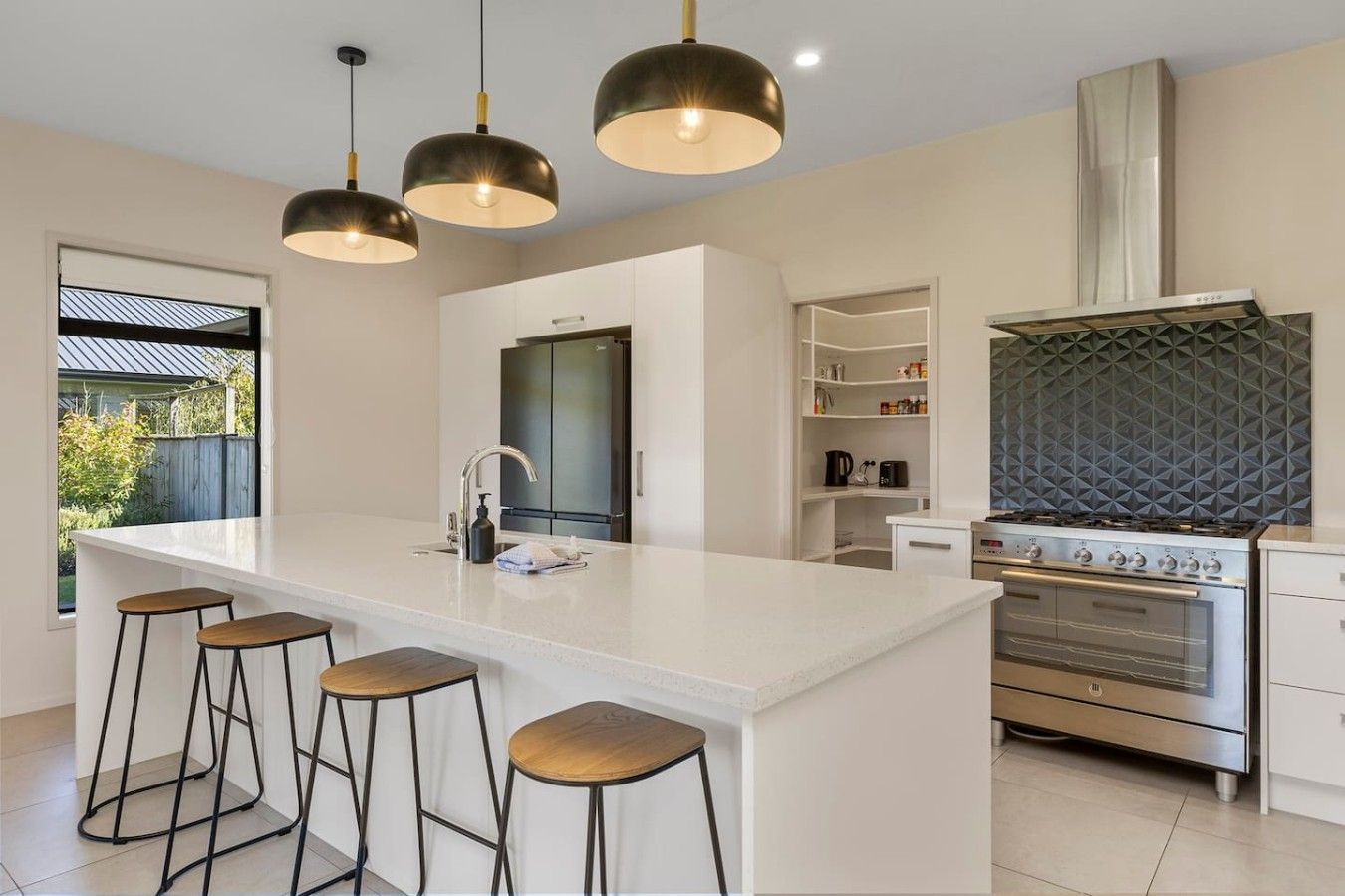 A modern kitchen with a central island, bar stools, pendant lighting, a built-in oven, and a backsplash with a geometric pattern.