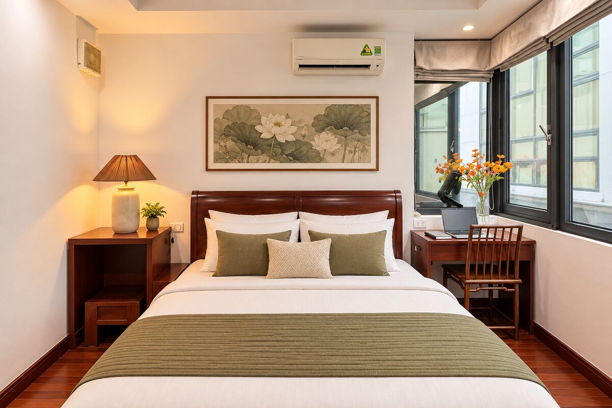 A hotel room with a bed, bedside table with a lamp, a desk with a laptop, a window with curtains, and a framed floral artwork on the wall.