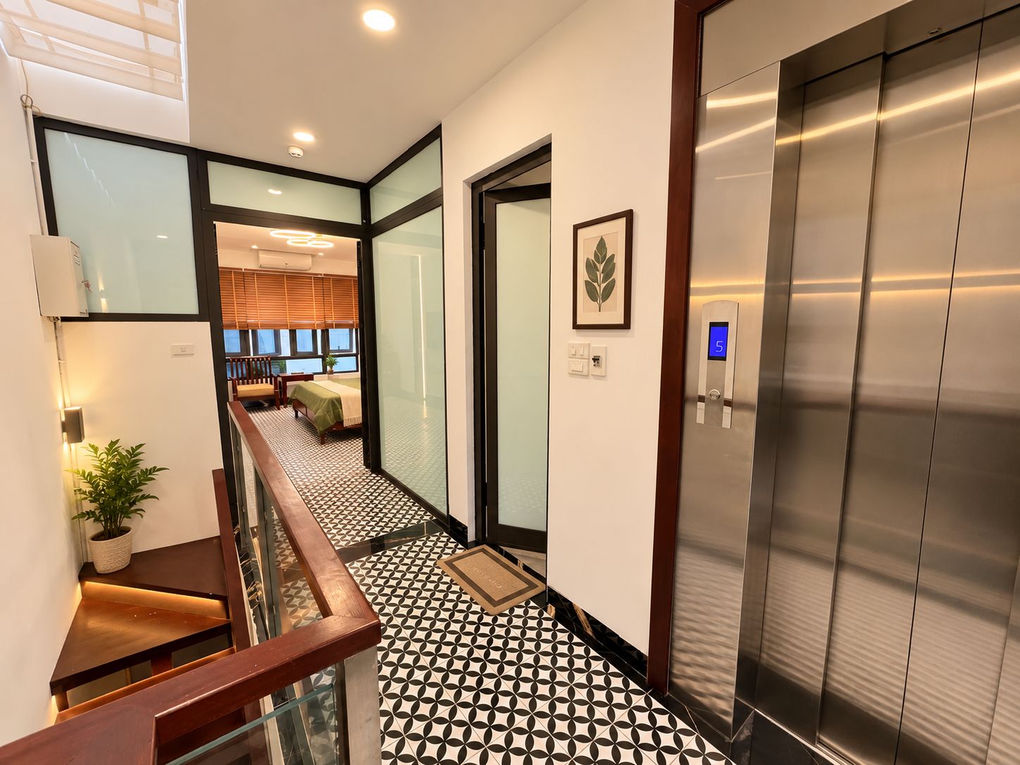 Eine moderne Hotelhalle mit einem geometrisch gemusterten Boden, einer Treppe, einem Aufzug und Blick in einen Raum mit einem Bett und Fenstern.