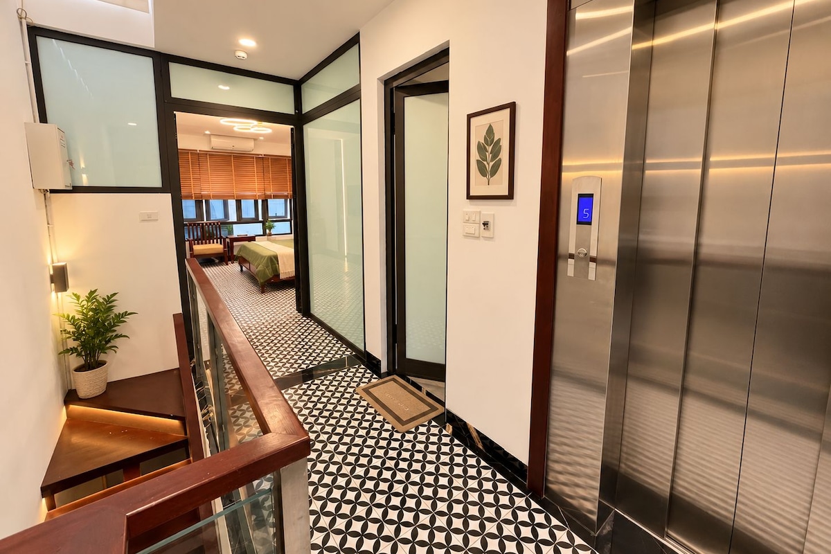 A modern hotel lobby with a geometric patterned floor, a staircase, an elevator, and a view into a room with a bed and windows.
