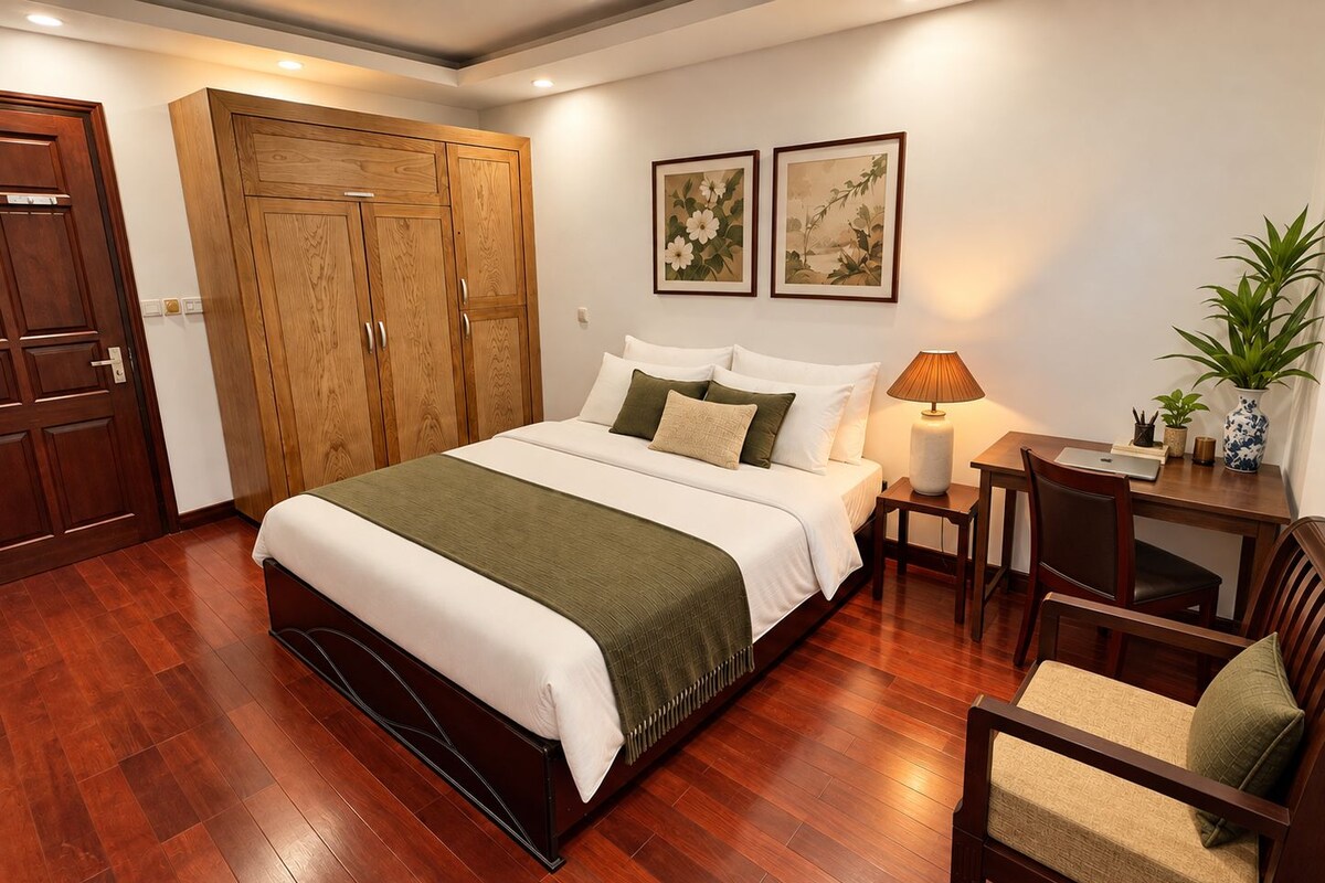 A bedroom with a large bed, wooden wardrobe, two framed artworks, a desk with a chair, and a potted plant.