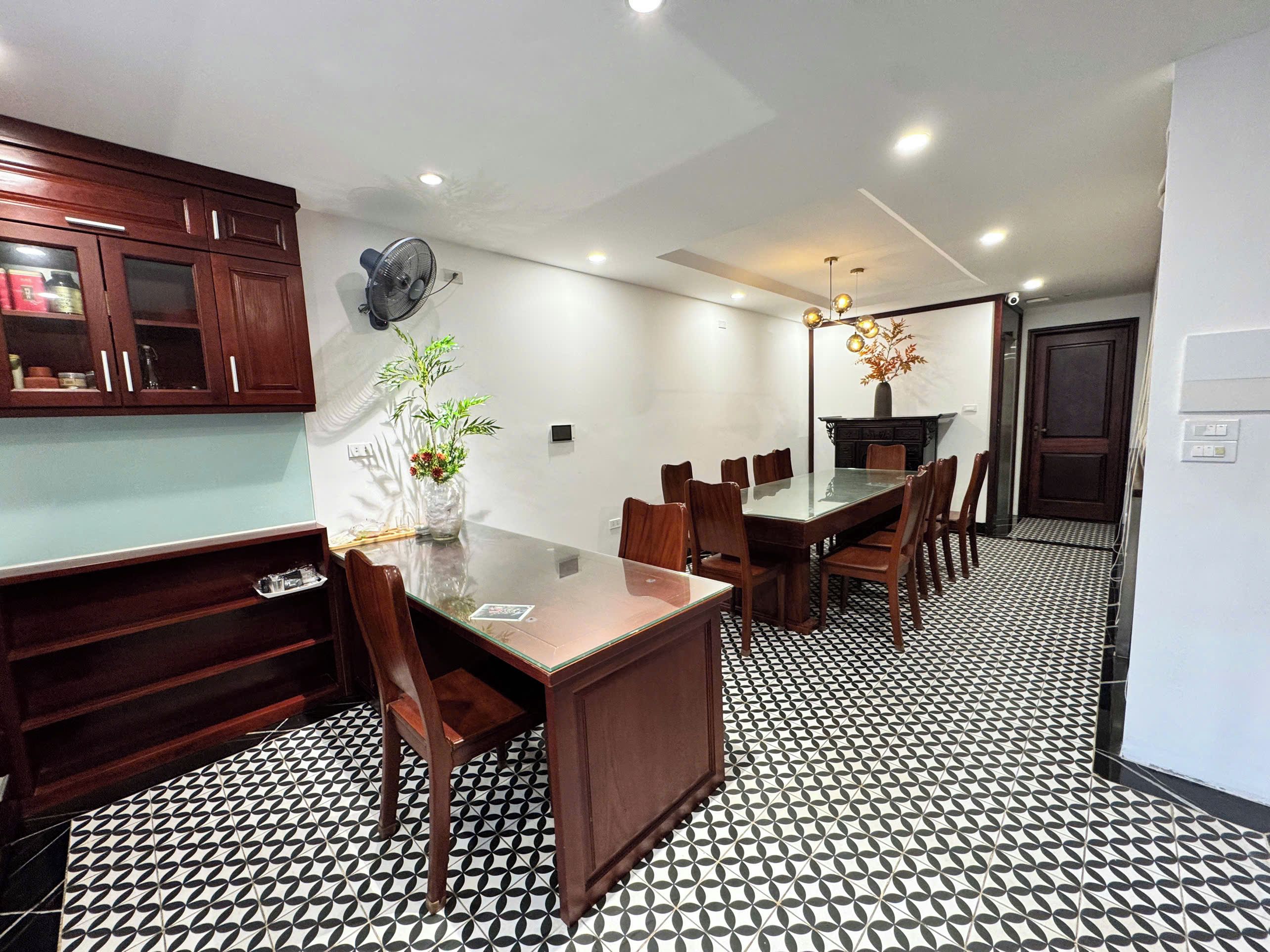 A dining area with a glass-top table, wooden chairs, a patterned floor, and a dark wooden cabinet.