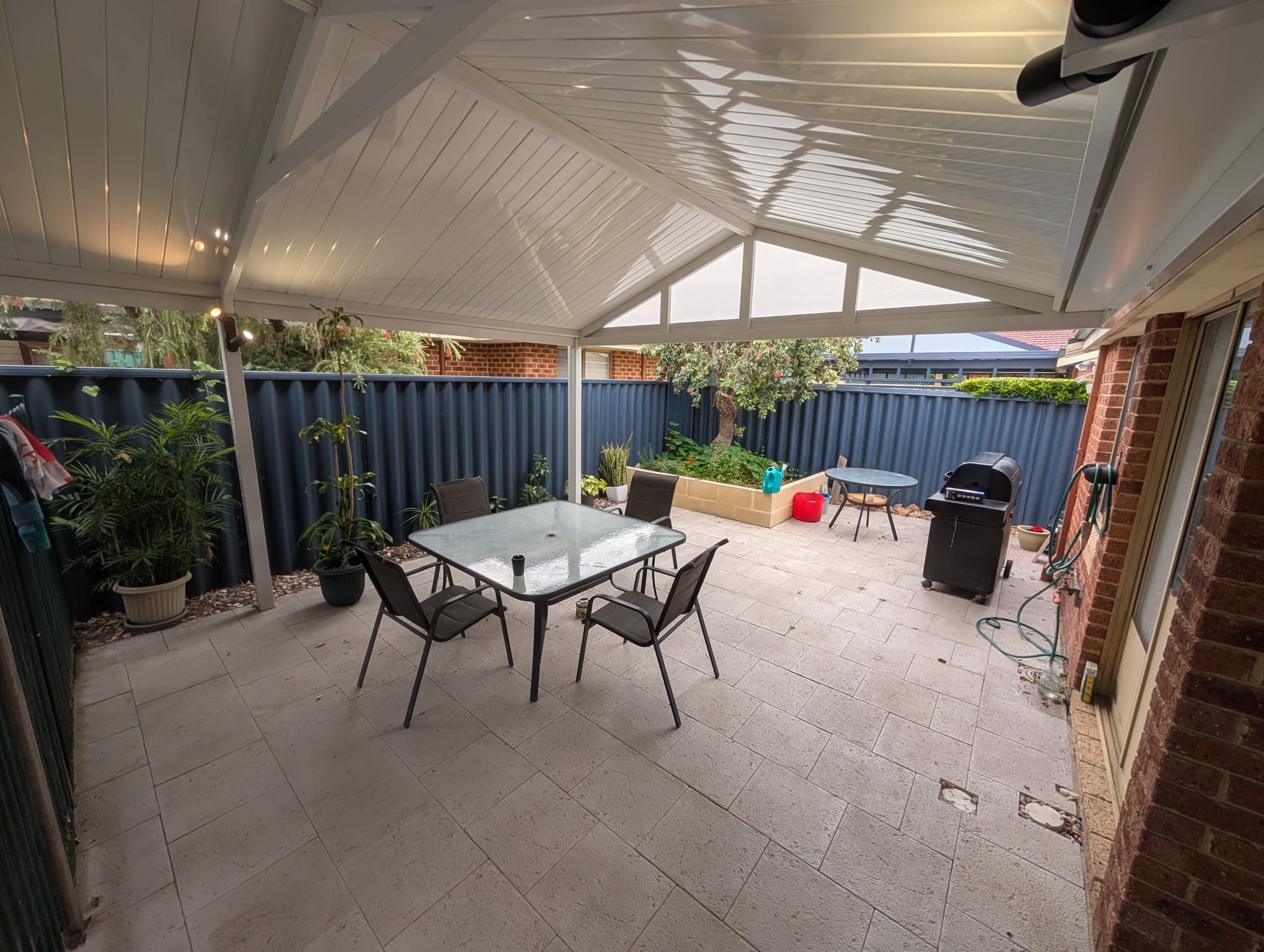 A covered patio area with a table and chairs, a barbecue grill, and potted plants.