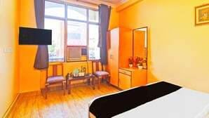 Quarto com cama, escrivaninha, duas cadeiras, televisão e janela com cortinas.