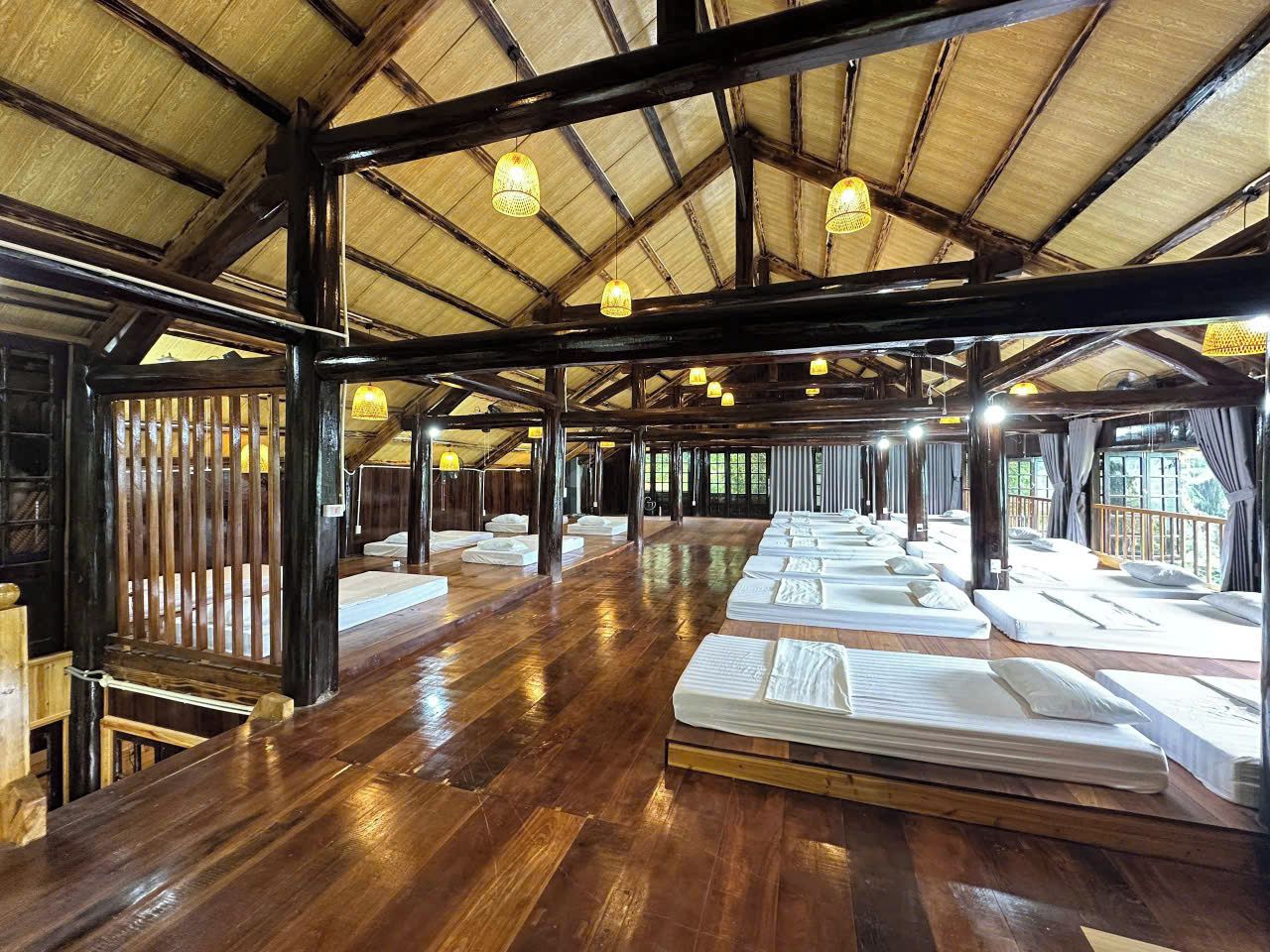 Un salón amplio con varias camas dispuestas en filas, vigas de madera y luces colgantes.