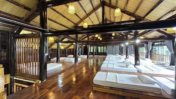 Un salón amplio con varias camas dispuestas en filas, vigas de madera y luces colgantes.