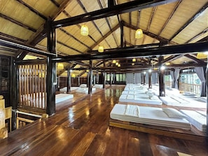 A large room with multiple beds arranged in rows, wooden beams, and hanging lights.