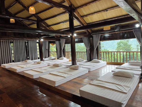 A room with multiple beds arranged in rows, wooden flooring, and a view of greenery outside.