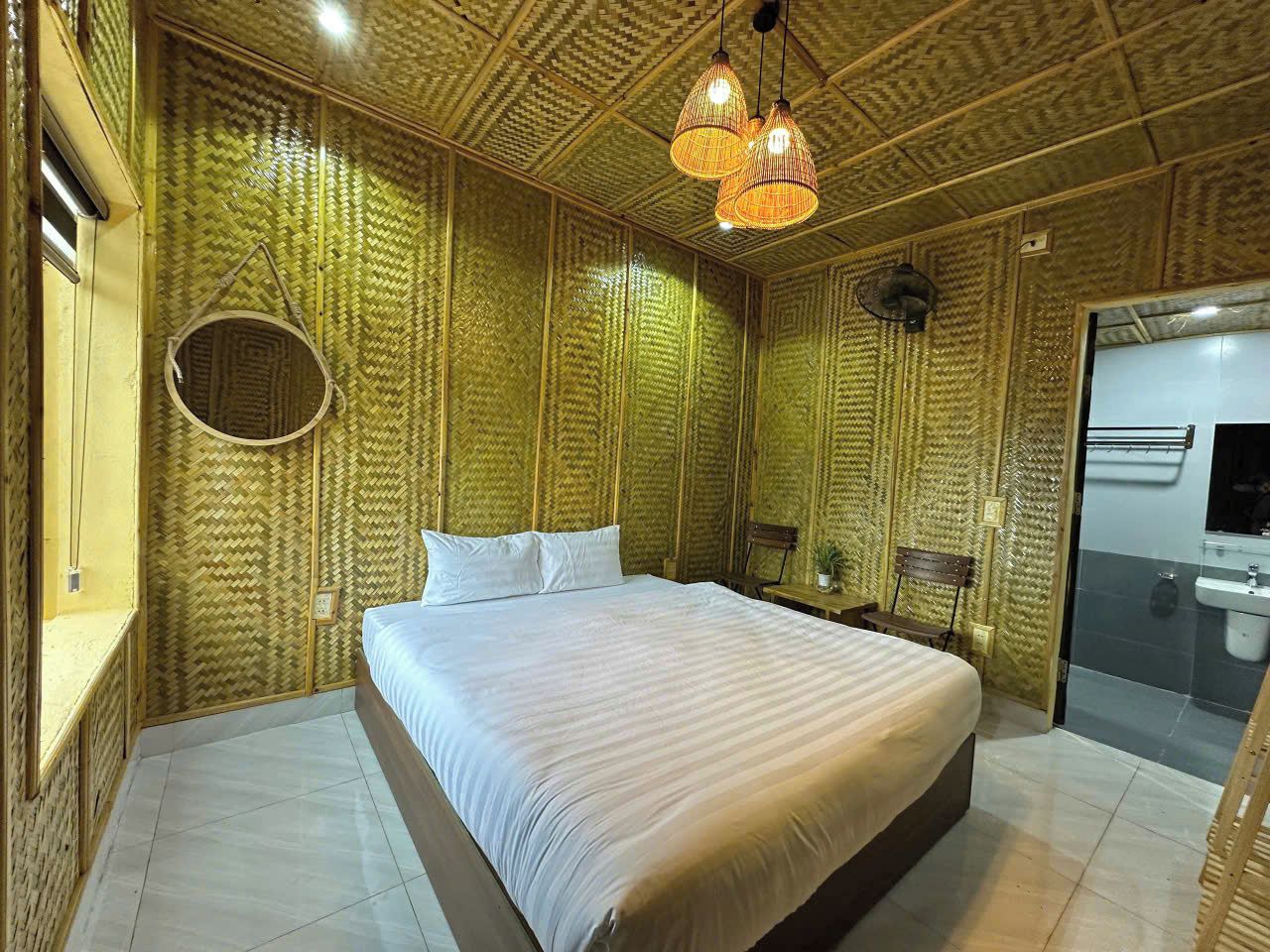 A room with a large bed, a round mirror, a wooden bench, and a bathroom with a sink and mirror.