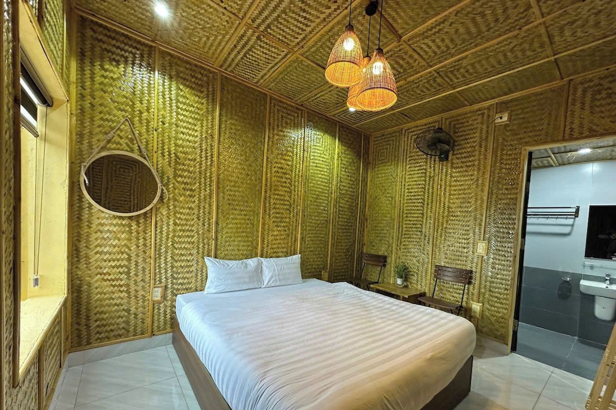 Una habitación con una cama grande, un espejo redondo, un banco de madera y un baño con lavamanos y espejo.