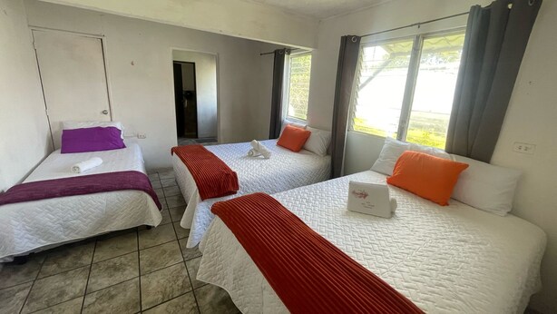 A hotel room with three beds, each with a red blanket, white pillows, and a white quilted bedspread. There is a window with curtains, a door, and a small table with a white box on it. - CASA AZALEA (Ciudad de Guatemala)