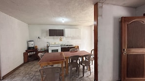 Una cocina compacta con armarios blancos, estufa y lavamanos. Hay una mesa de comedor con sillas y una puerta de madera.