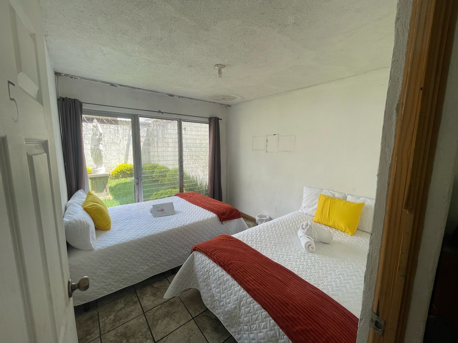 A hotel room with two beds, a window with curtains, and a view of greenery outside.