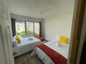 A hotel room with two beds, a window with curtains, and a view of greenery outside.