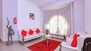 A living room with a white sofa, red cushions, a glass coffee table, a statue, a Buddha statue, and a fire extinguisher on the wall.
