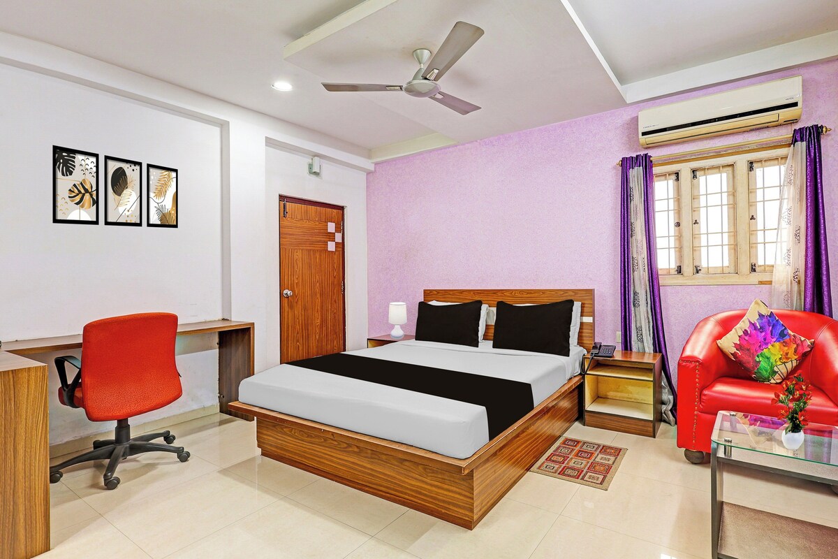 Une chambre d’hôtel avec un lit, un bureau, une chaise, un canapé rouge, une fenêtre avec des rideaux et un ventilateur de plafond.