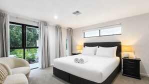 Ein Hotelzimmer mit einem großen Bett, zwei Nachttischen, einem Sessel und einem Fenster mit Vorhängen.