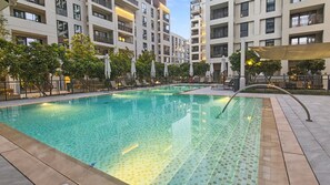 Ein Pool mit Fliesen umgeben, mit Liegestühlen und Sonnenschirmen, im Hintergrund ein mehrgeschossiges Gebäude.