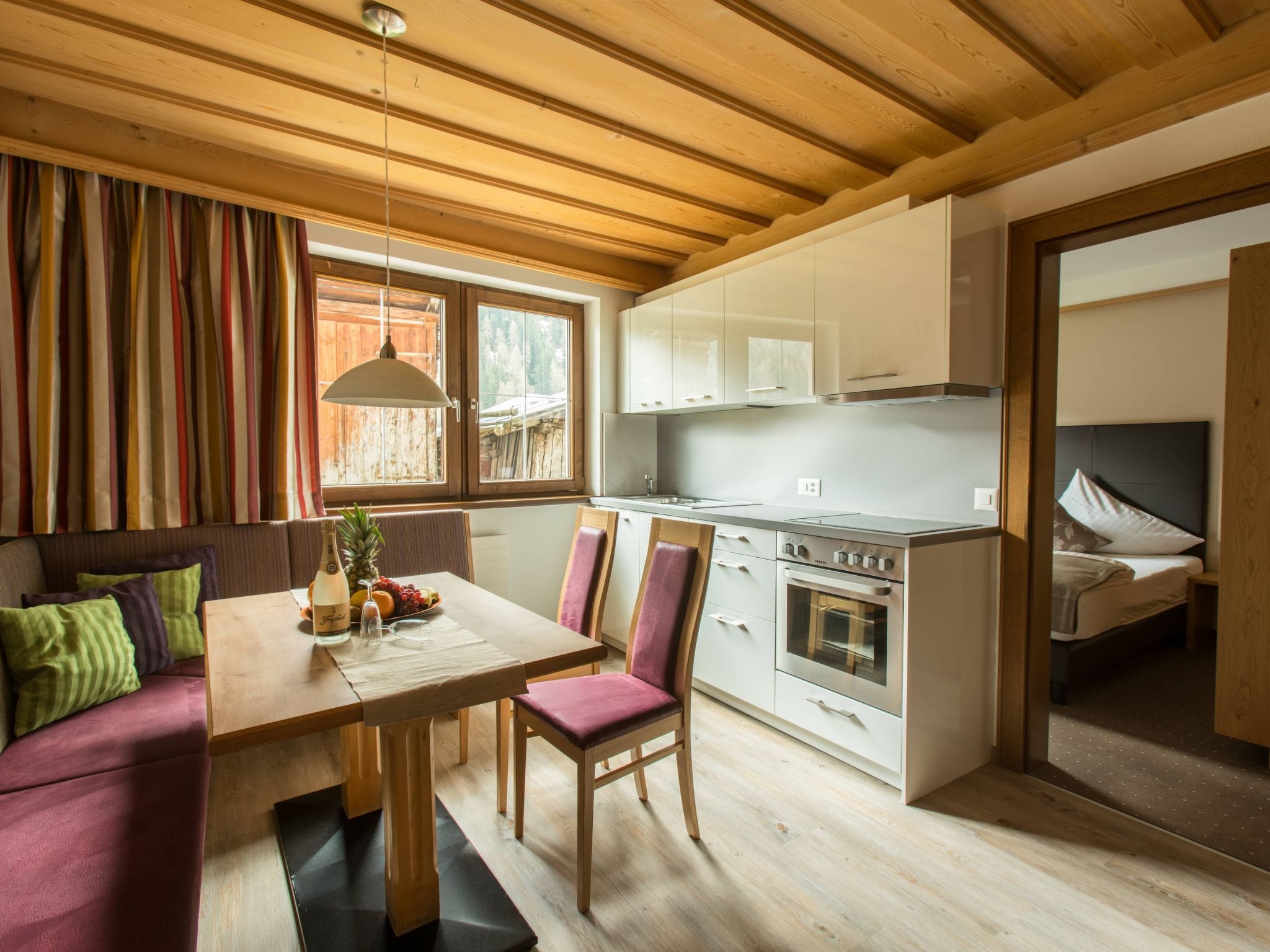 A cozy kitchen with wooden beams, a dining table with chairs, and a view of a bedroom.