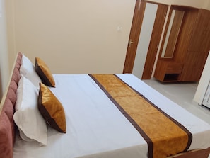 A neatly made bed with a brown accent, a wooden wardrobe, and a door in a hotel room. - Shivansi Residency (Varanasi)