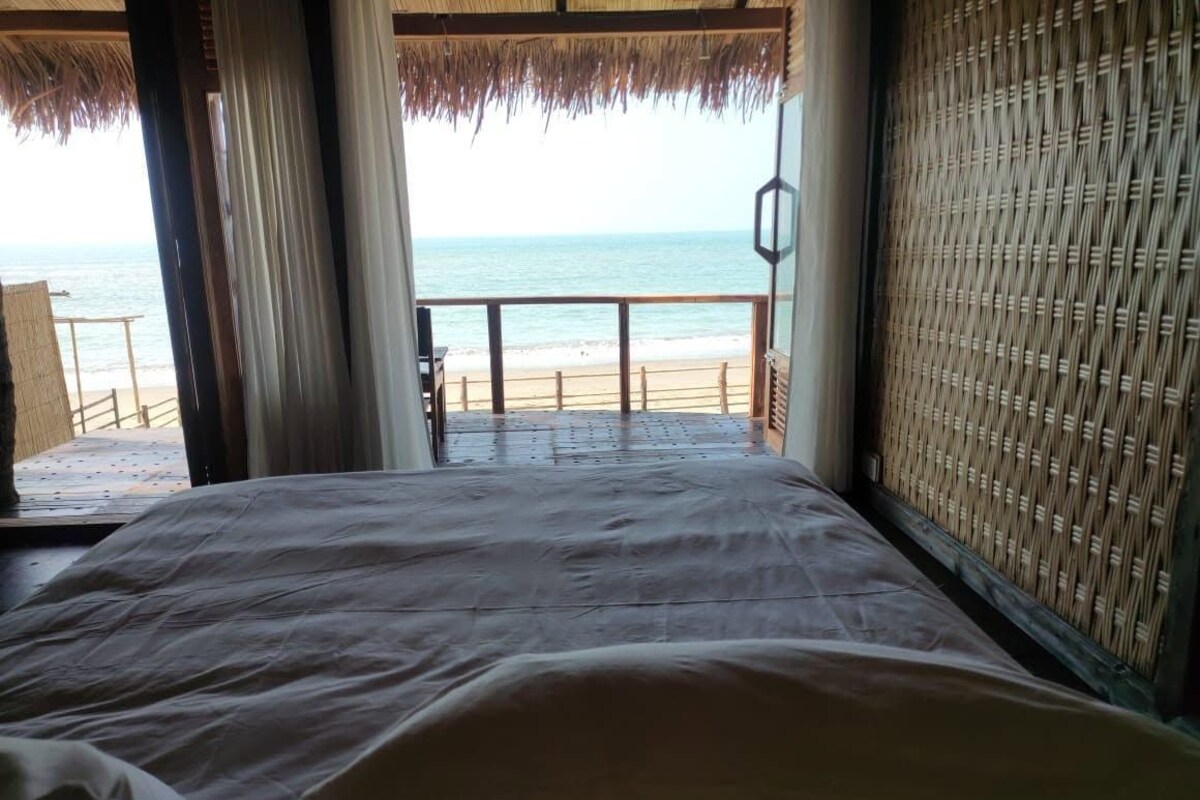 A room with a bed, a view of the beach, and a thatched ceiling.