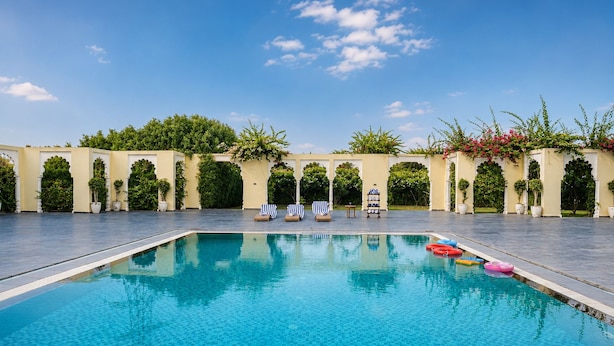 A swimming pool with clear blue water, surrounded by a paved deck, and flanked by greenery and architectural arches. - Beelwa Palace  (Jaipur)