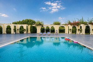 Una piscina con acqua cristallina, circondata da un solarium in pietra e fiancheggiata da vegetazione e archi architettonici.