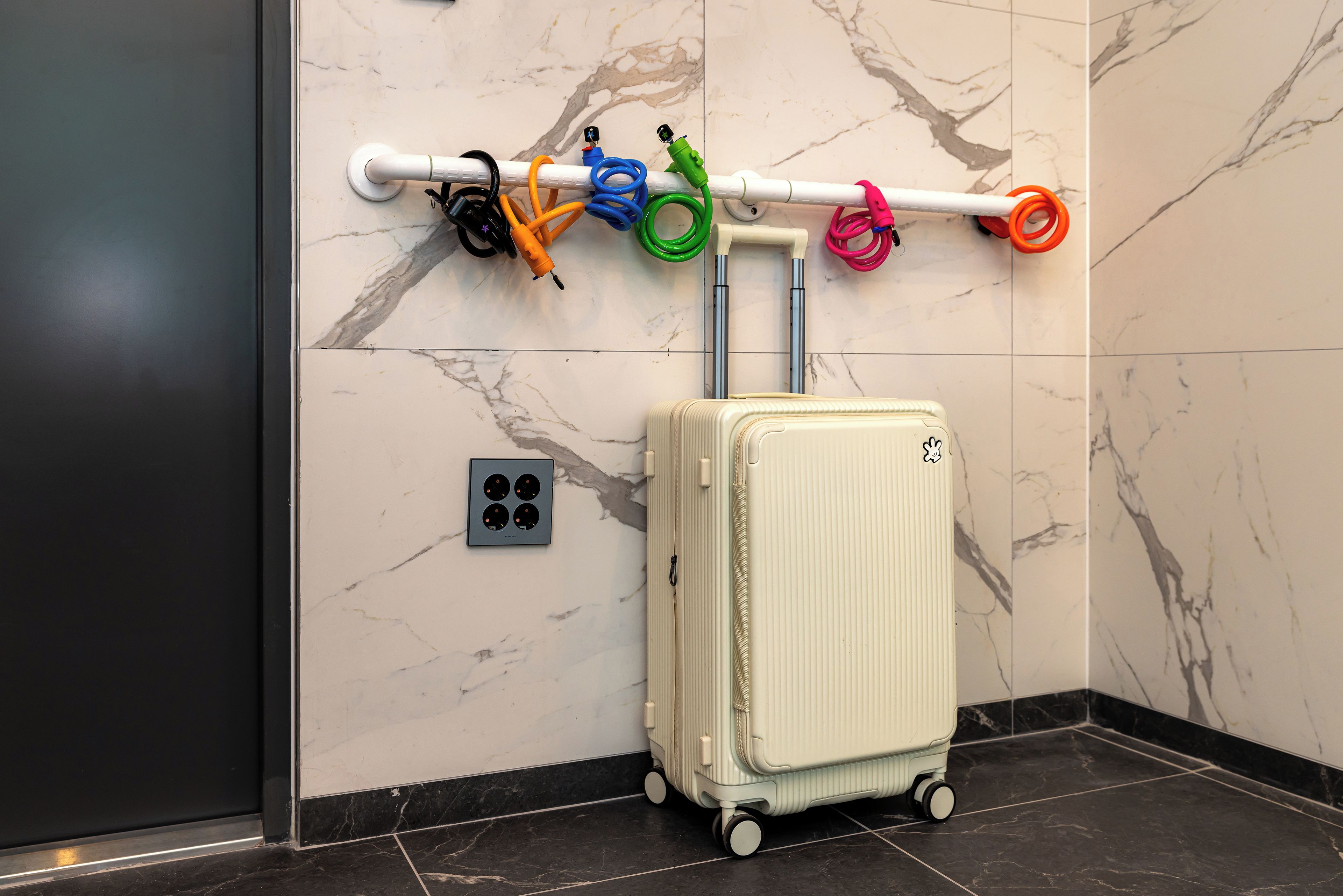A wall-mounted luggage rack with multiple colored straps, a power outlet, and a white suitcase with wheels.