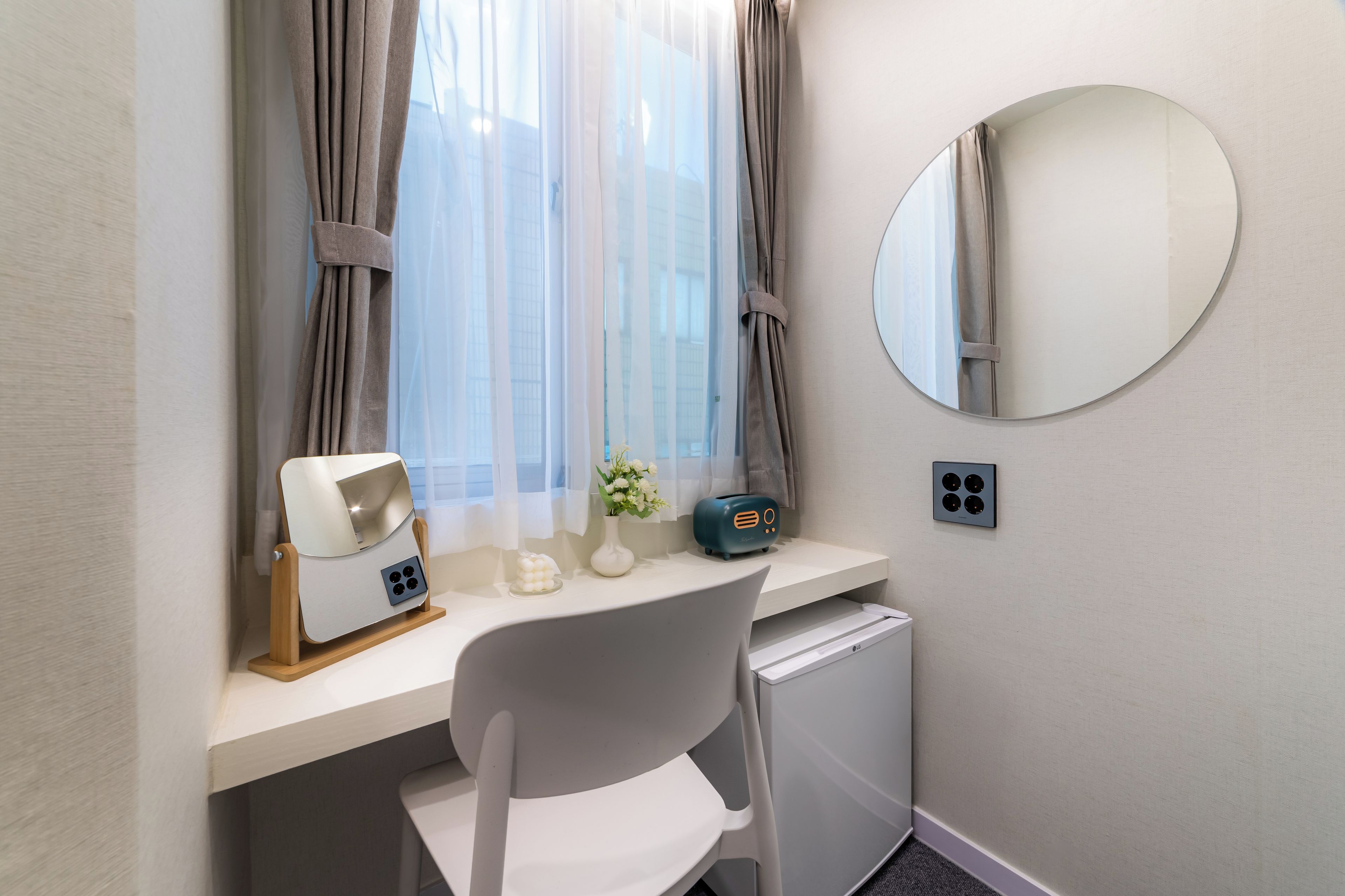 A hotel room with a desk, a chair, a mirror, a small table with a lamp, a radio, and a compact refrigerator.